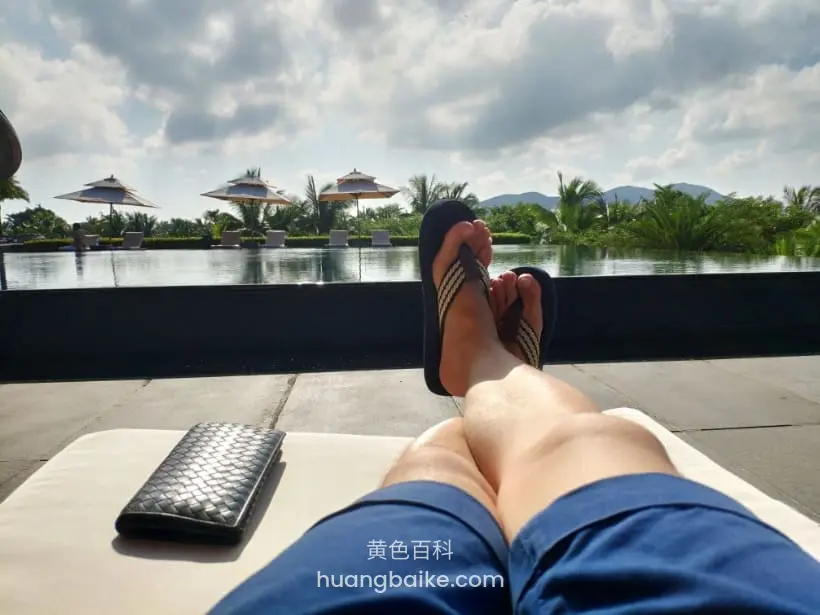 度假酒店躺椅上翘腿,脚边放编织包,身后是湖水和亭子