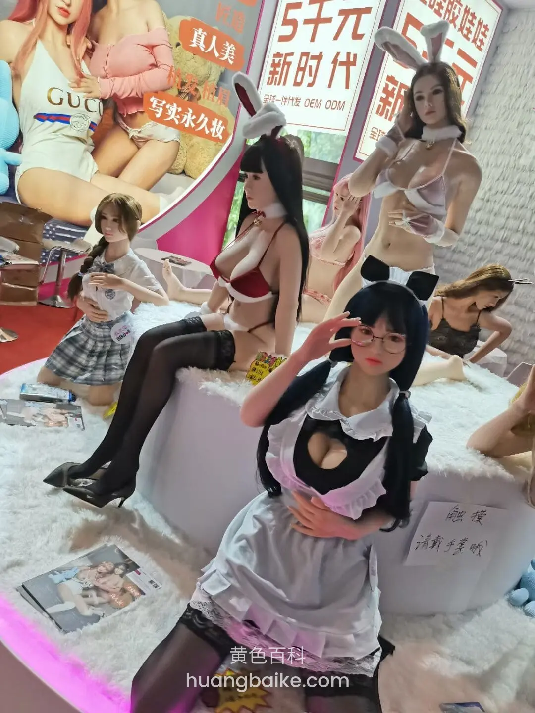 展台白色平台前坐着比V手势,身后一排情趣娃娃和海报