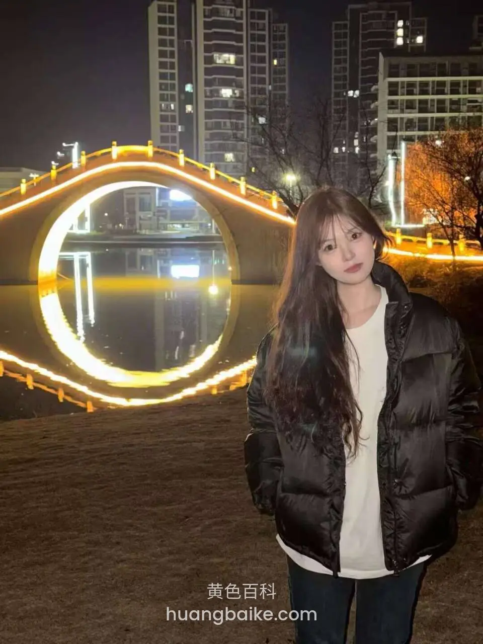 夜晚河边拱桥前站立，身后灯光倒影在水面