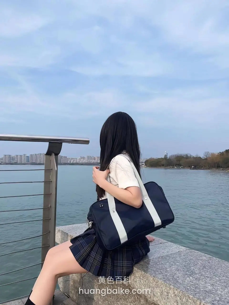 坐在湖边栏杆上背对镜头，手抱蓝色书包，远处城市天际线