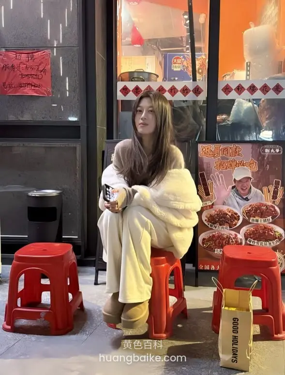 街边小店门口坐红凳 手里拿着手机 旁边有麻辣烫招牌和GOOD MOOD纸袋