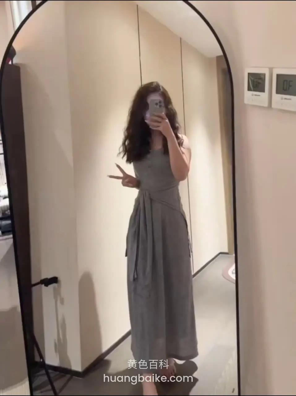 长发女人穿着灰色连衣裙站在拱形镜子前自拍，左手比剪刀手，赤脚站在地上