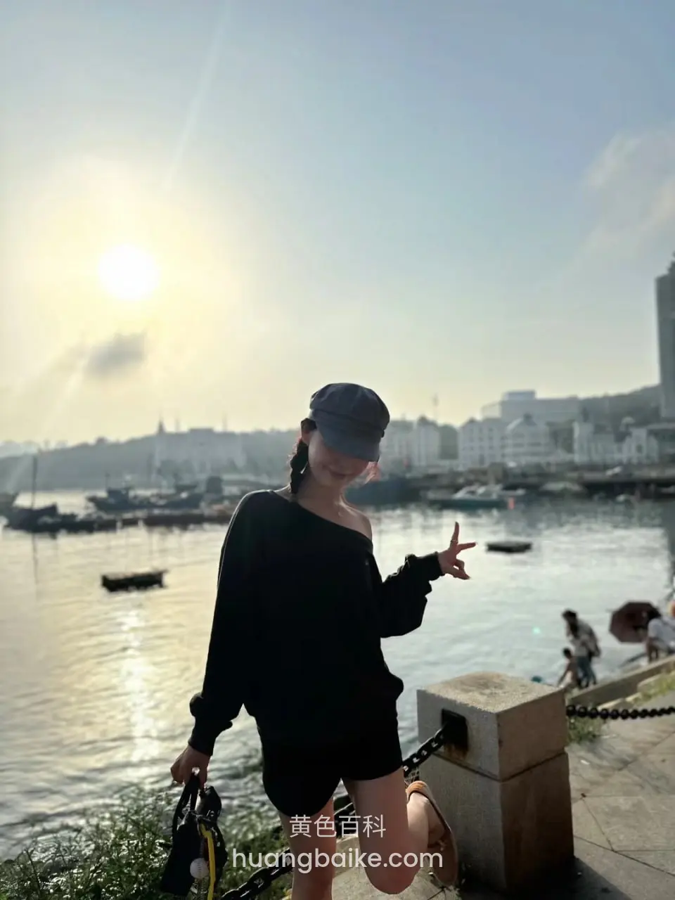 港口边单膝跪地做剪刀手，身后船只和夕阳