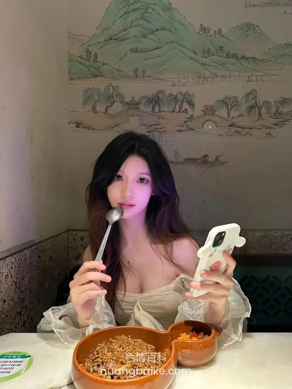 餐厅桌前拿着勺子吃饭，左手举着白色手机