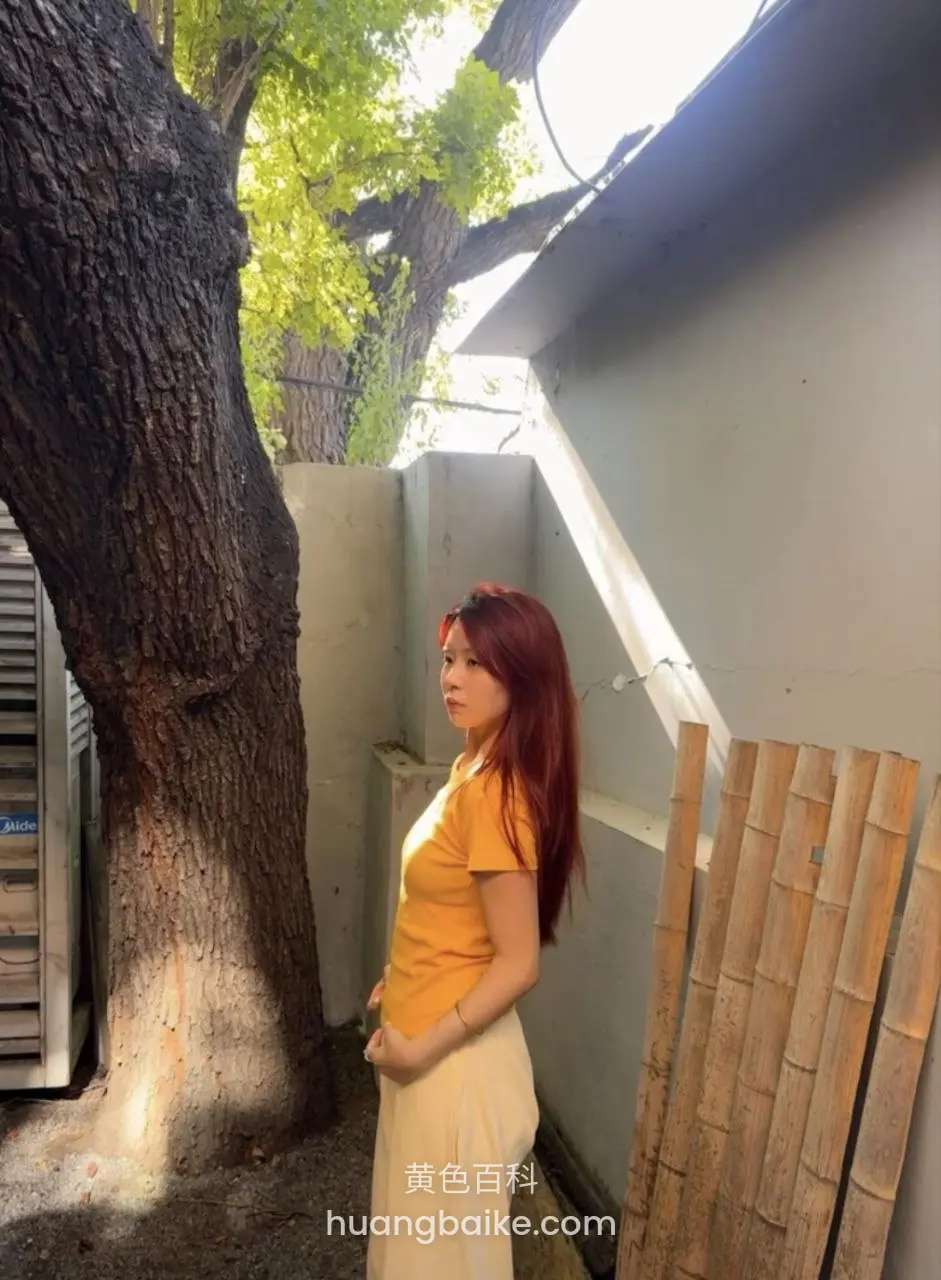 红发女孩穿着黄色T恤和白裙站在树边，侧身看向镜头，阳光洒在身上