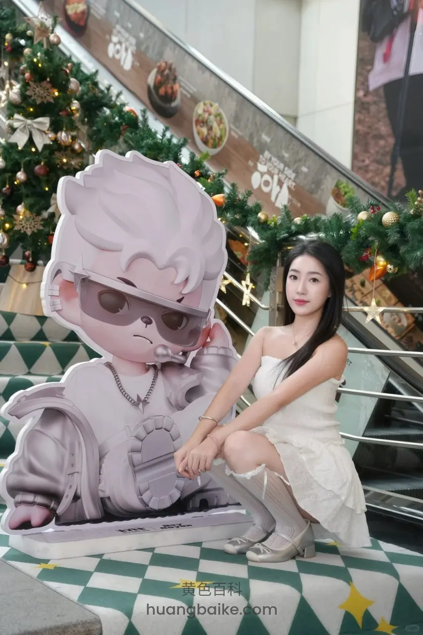 商场圣诞树旁蹲下，右手搭着紫色卡通人偶