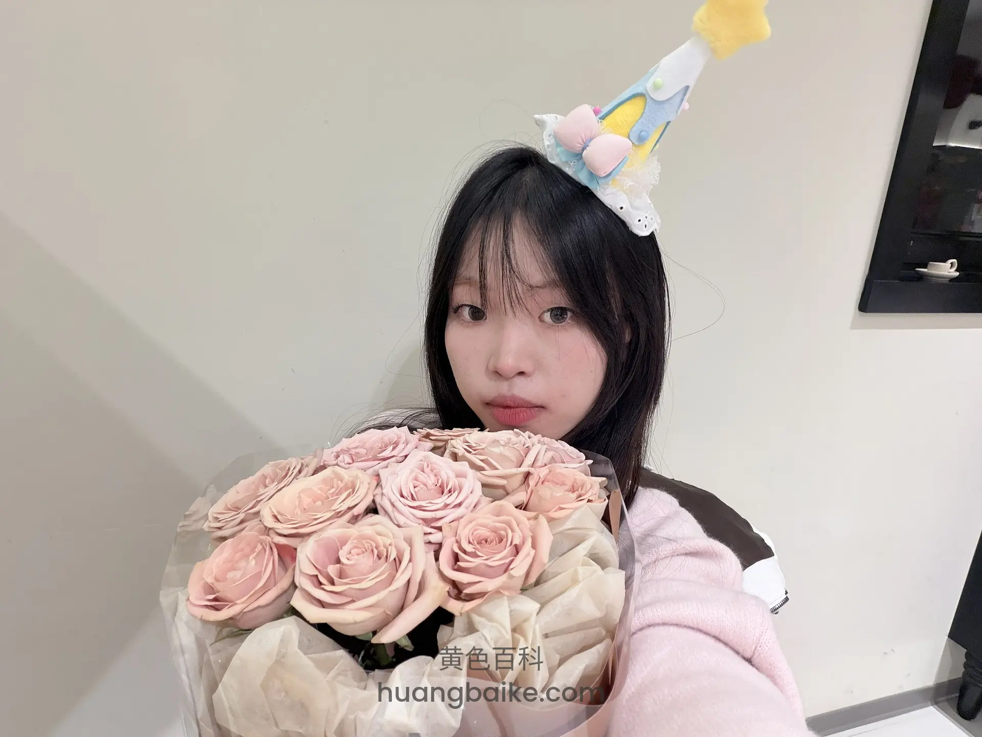 室内举着粉玫瑰花束，头顶生日帽