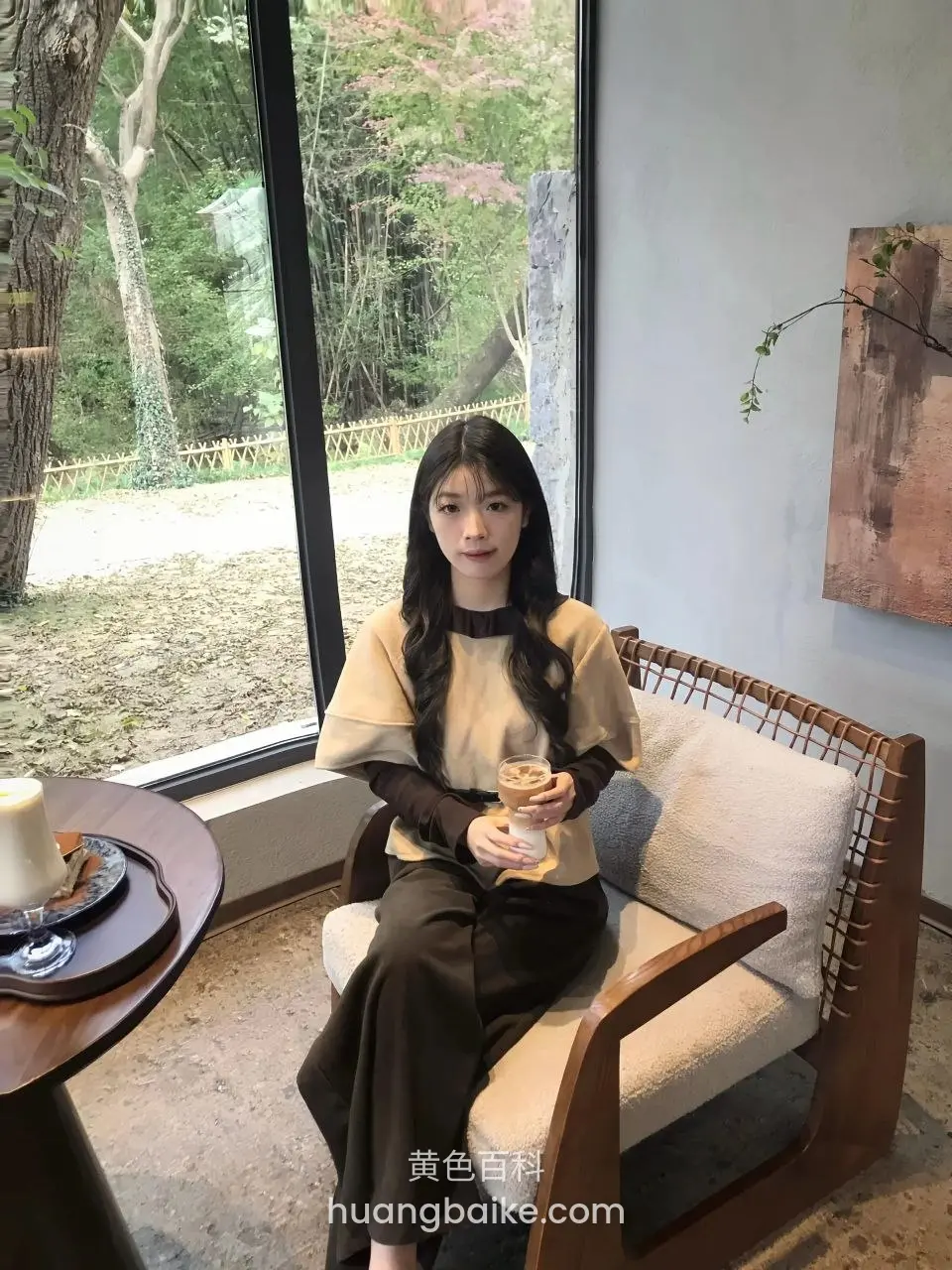 拿着咖啡杯坐在窗边椅子上，身后是树木