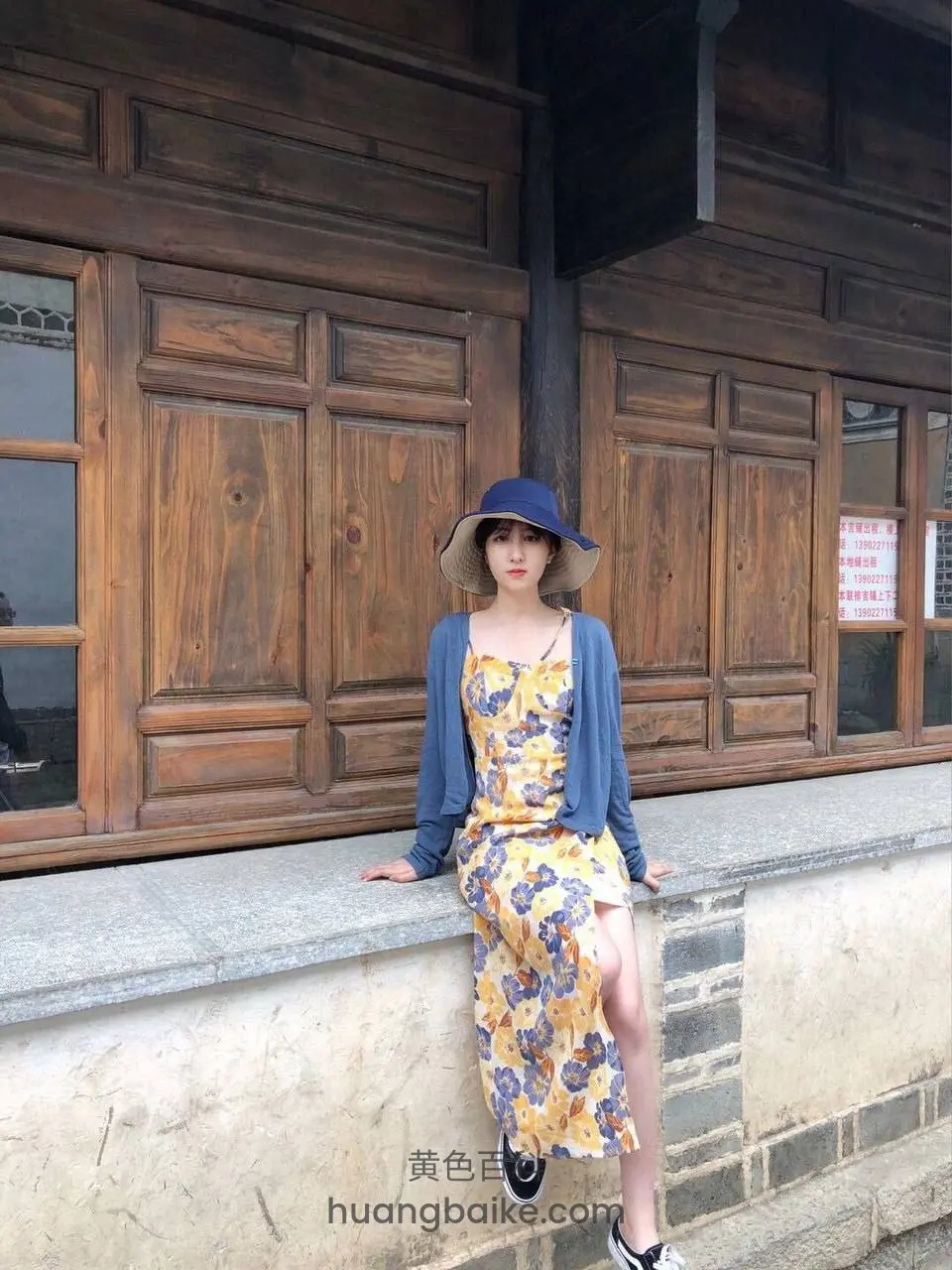 古建筑木门前靠石栏坐着，穿花裙戴帽