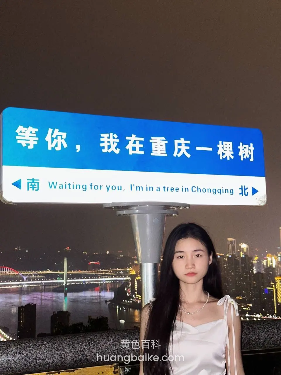 夜晚江边路牌前站立，身后是重庆灯光夜景